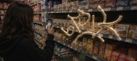 Someone photographs a glowing 3D 'GLYF' sculpture anchored among supermarket shelves.